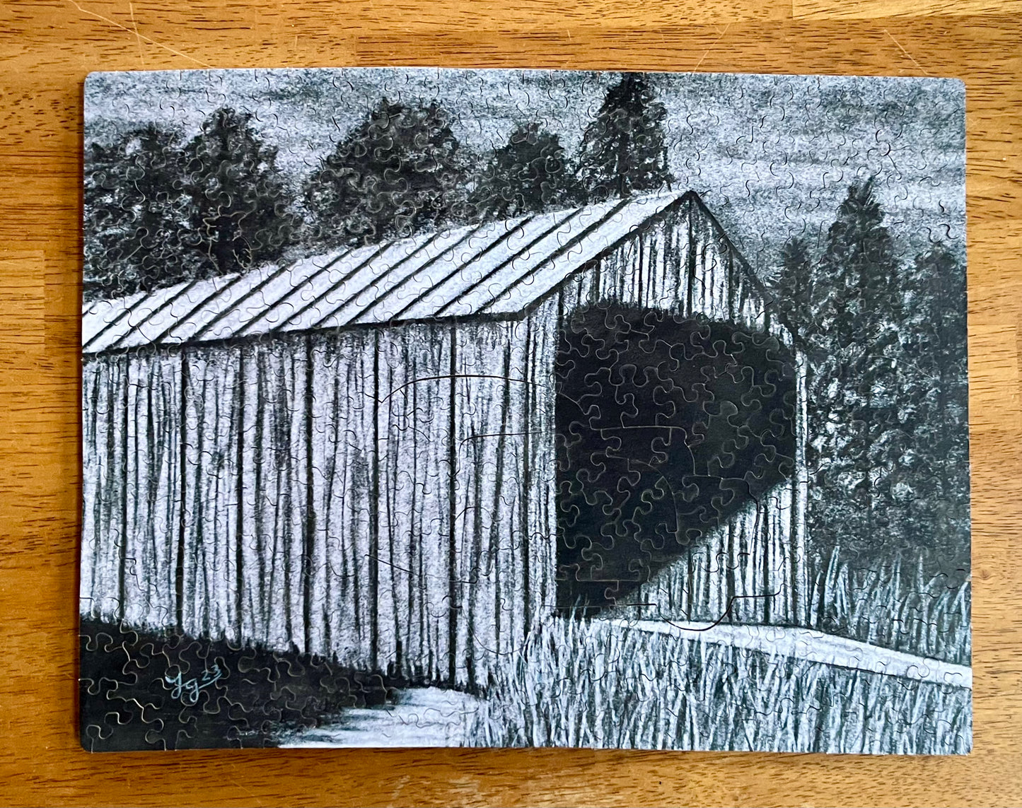 This image displays the "Covered Bridge Puzzle," a handcrafted jigsaw puzzle that features a charcoal drawing of a covered bridge by artist Jenell Green. The puzzle is composed of multiple pieces, some of which are uniquely shaped whimsies, adding a distinctive touch to each puzzle's configuration.